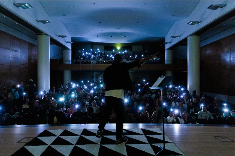 A person performing on a dark stage in front of a crowd holding up lit phones