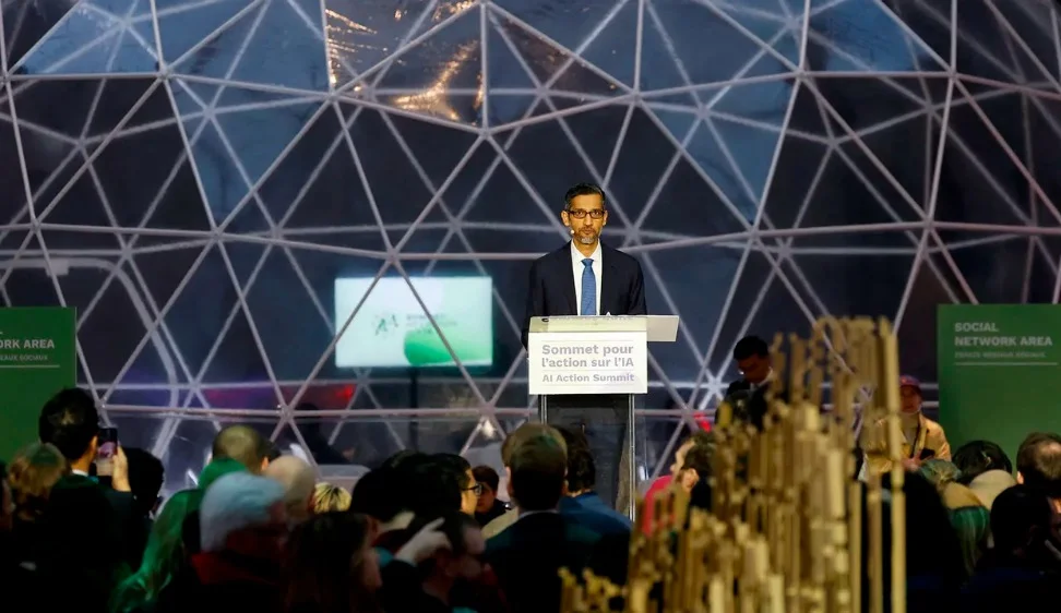 A photo of Google CEO Sundar Pichai standing on a stage at the AI Action Summit, giving a speech behind a podium.