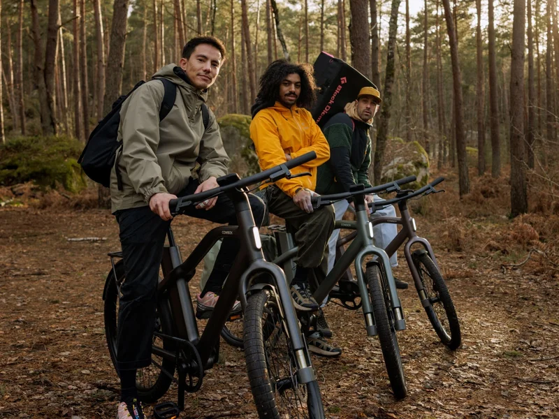 Three people with Cowboy bikes standing on a wooded trail. The group is posed casually, with trees and sunlight creating a natural backdrop. Each bike features the Cowboy branding and sleek design.