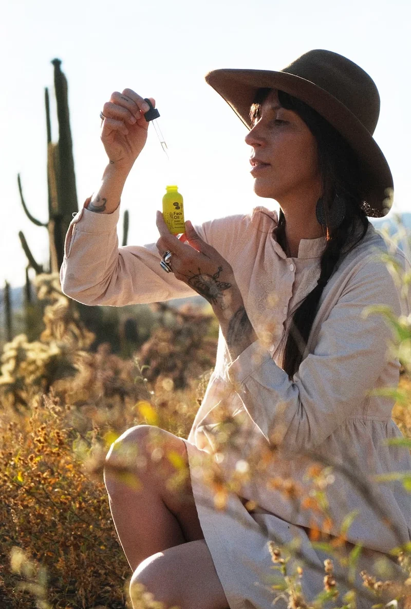 A person with a brown hat sits in a desert and puts face oil on