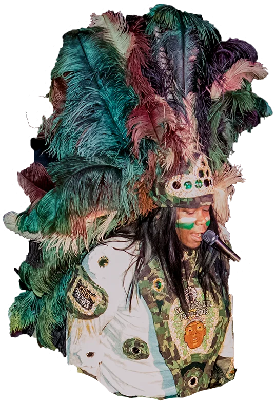 Performing artist in vibrant traditional feathered headdress and camouflage outfit singing into a microphone.