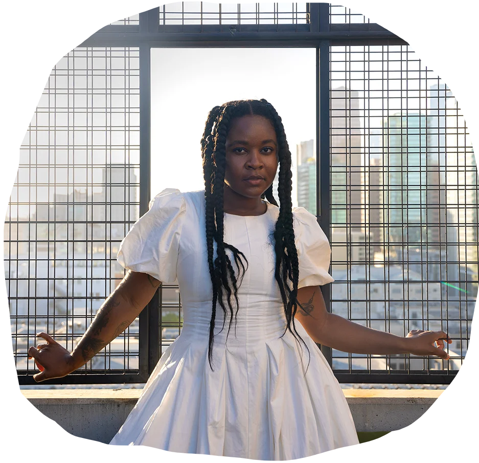 Elegant woman with braided hair and tattoos wearing a white puff-sleeve dress posing confidently on an urban rooftop with a cityscape in the background.