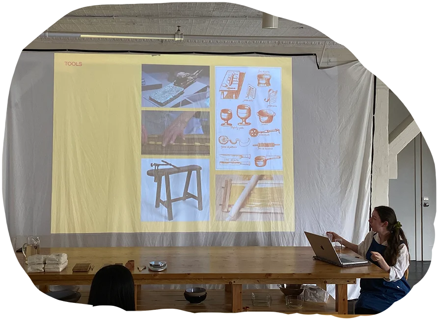 Presenter at a casual workshop with a laptop, speaking beside a screen displaying a collage of traditional tools.