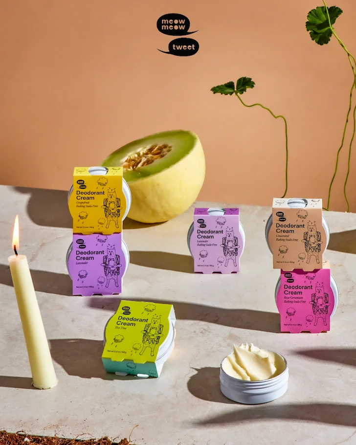 A variety of cream deodorants sits on a tan background with a burning taper candle and half of a melon