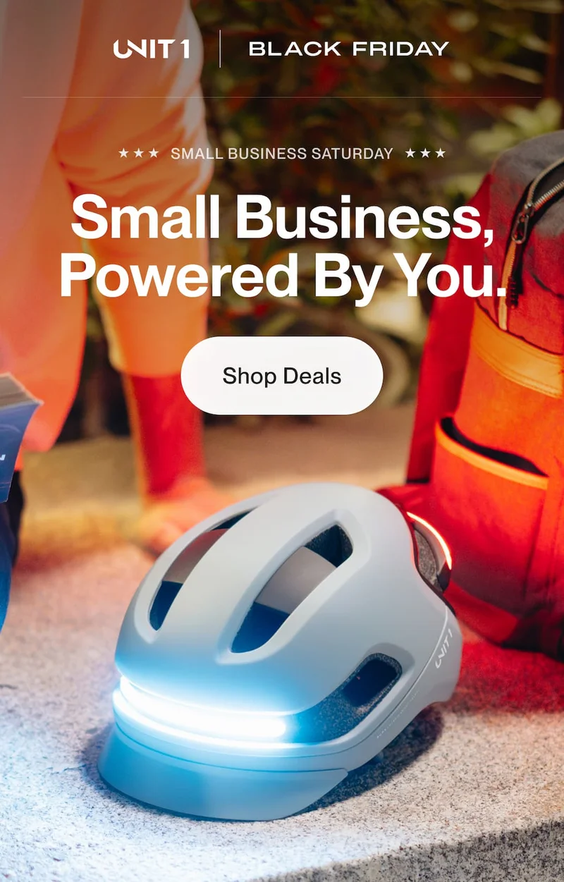 UNIT 1 smart helmet with front and rear lights illuminated, shown outdoors during Small Business Saturday with a Shop Deals call to action.