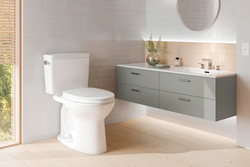 A TOTO Drake Toilet in a sunlit bathroom near a tall window and sage vanity