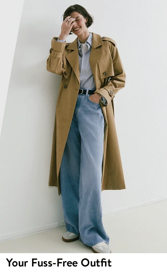 A woman wearing a tan trench coat, button-up shirt, wide-leg jeans and white sneakers.