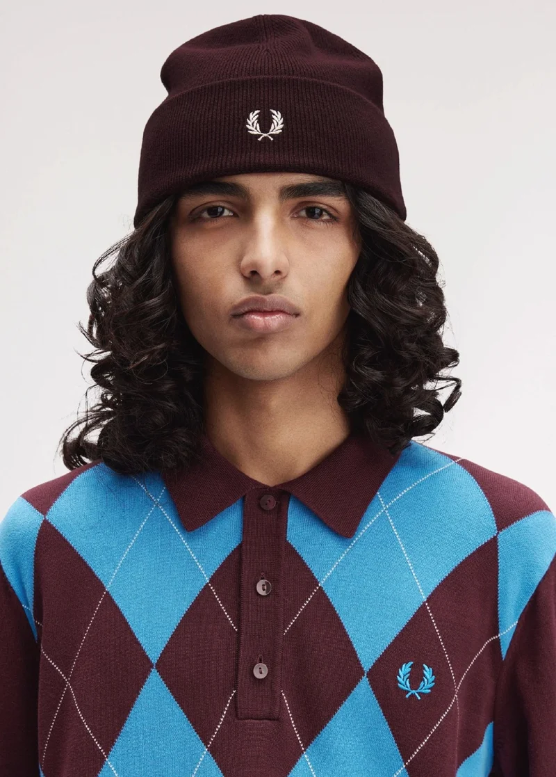 Male model with shoulder length wavy hair wears a knitted argyle long-sleeved polo shirt in bright blue and Oxblood. He wears a matching Oxblood knitted beanie with a white embroidered laurel wreath logo in the centre.