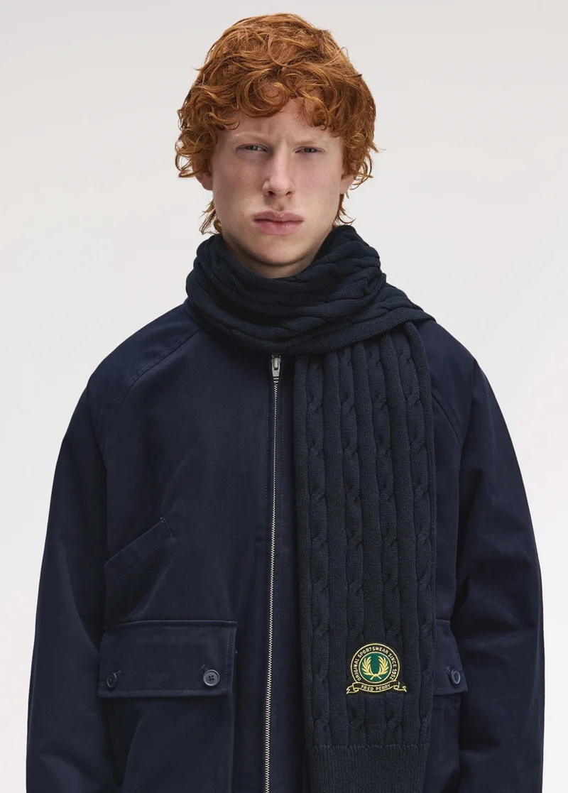 A male model resembling a young Prince Harry with wavy ginger hair wears a navy jacket with a cosy cable knit navy scarf draped around his neck. The scarf features a circular 'club house' inspired Fred Perry logo at the end, reminiscent of a sporting team logo.