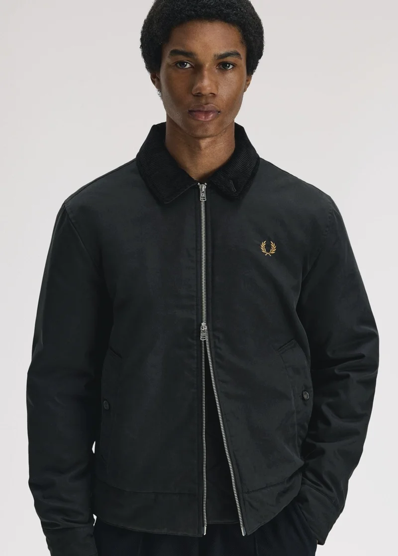 Black male model with black textured hair looks into the camera and stands nonchalantly with one hand in his pocket. He wears a black Caban jacket, with a silver zip, a light brown laurel wreath logo embroidered on the top left, and a corduroy collar, also in black.