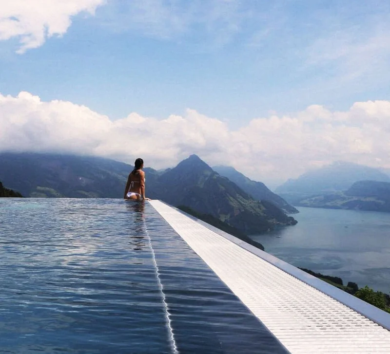 Image Description: A female traveler gazing at a faraway mountain range from an outdoor infinity pool at Hotel Villa Honegg, a hotel in Switzerland