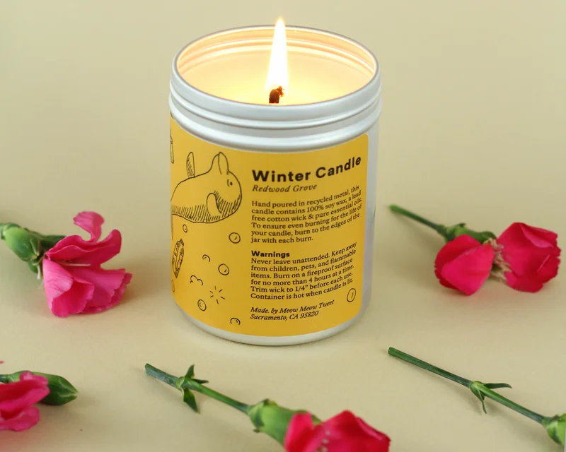 A lit candle in a metal jar with a yellow label with pink flowers strewn about it.
