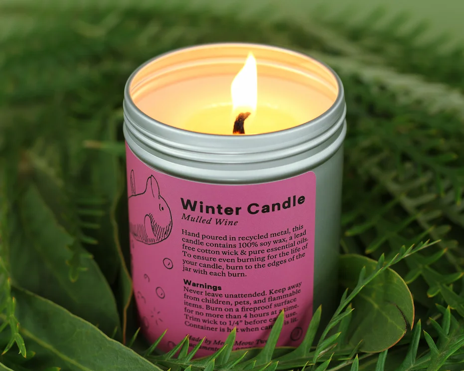 A lit candle in a metal jar with a pink label nestled in green plants.