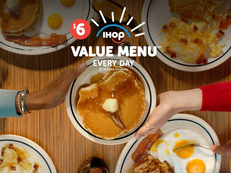 Two hands reach across a table and hold a plate of pancakes, topped with syrup and butter, from either side. Other plates of IHOP food are on the table below it.