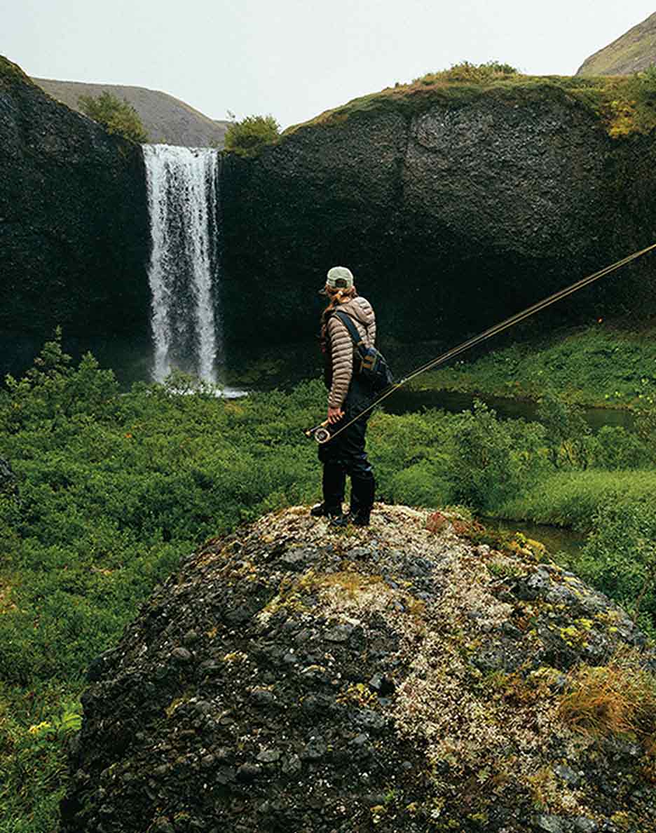 An angler stands on a rock overlooking a waterfall’s plunge pool.