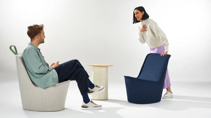 On the left is a man sitting in a white Pippin chair talking and facing a woman on the right standing over a dark blue Pippin chair, and a white stool in the background.