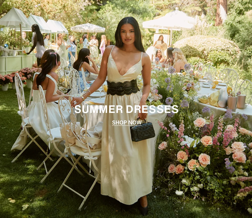 Summer Dresses: Elegant woman in cream plunge dress & corset belt at a lush garden party. Shop women's fashion collection now.