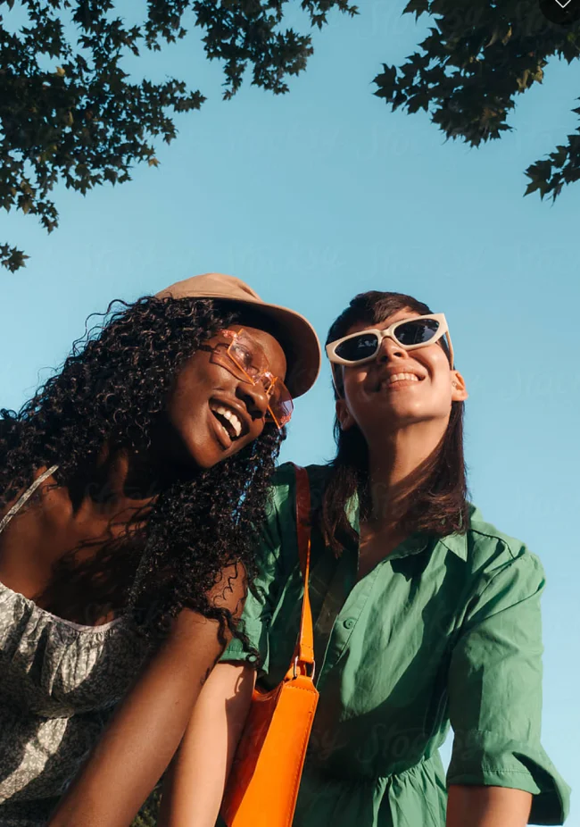 Two women smile while wearing sunglasses outside.