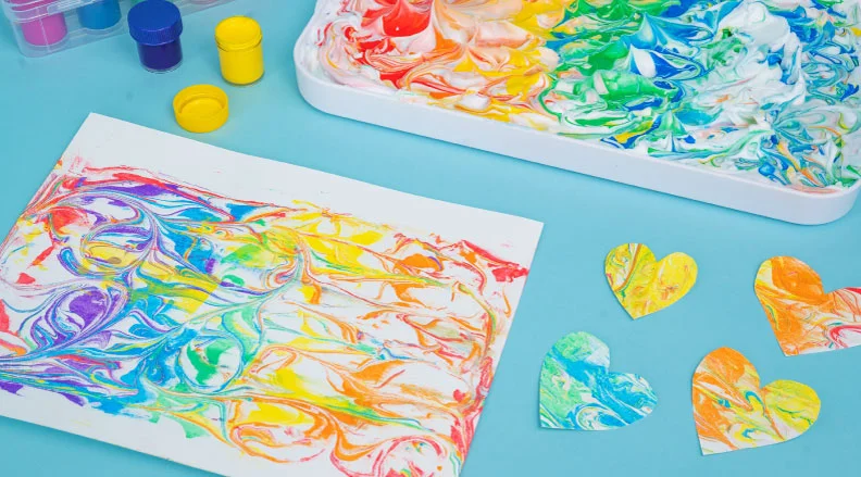 Colorful marbled paper with heart-shaped cutouts on a crafting tray with paint bottles around