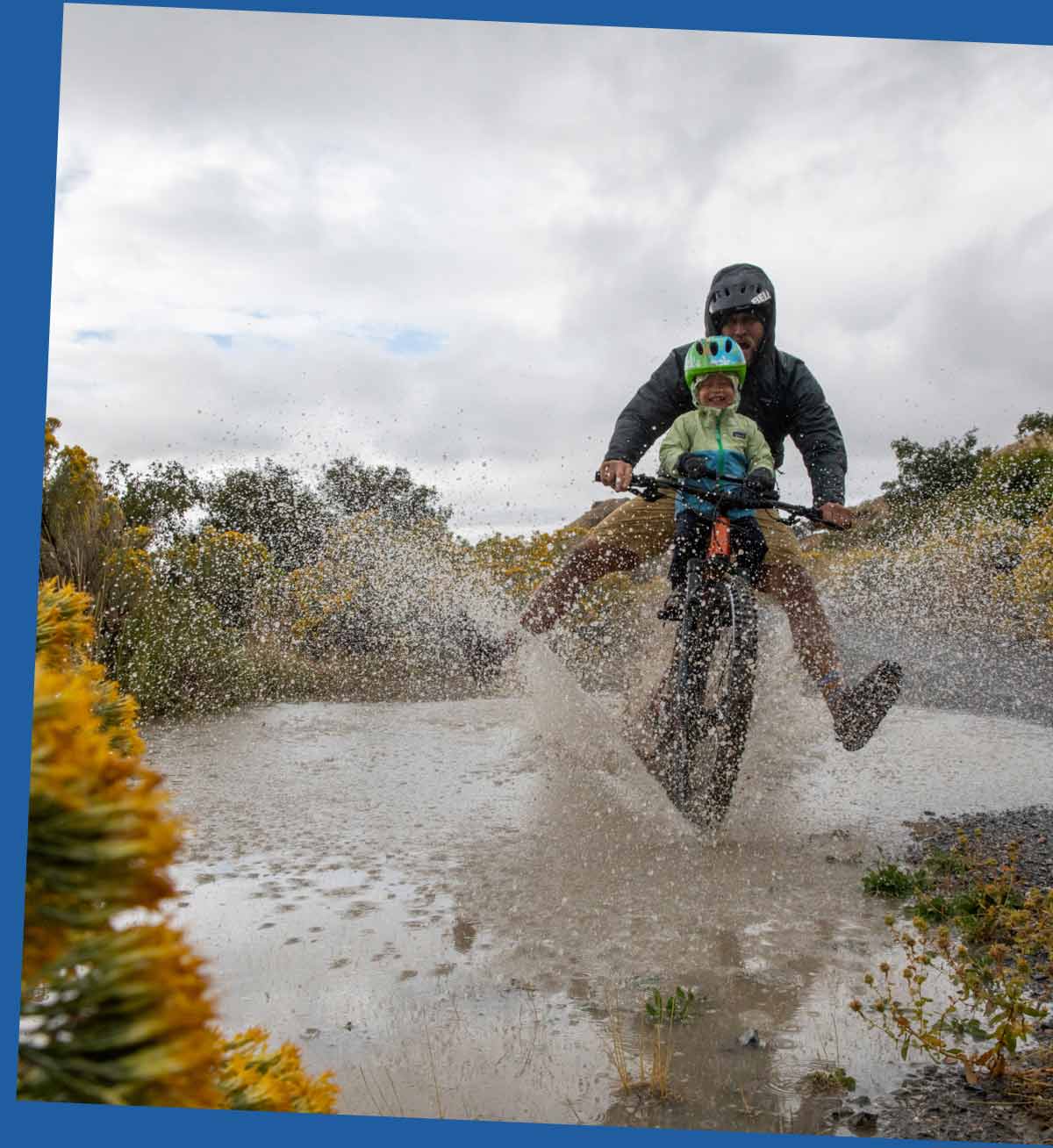 A parent and child ride a bike through a puddle making a big splash.