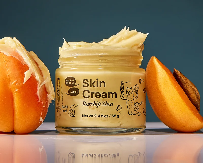 A glass jar with black line illustrations (cats holding morel mushrooms) stands open with skin cream swirled at the top. A couple of apricots lean up agains the jar with cream smeared on them