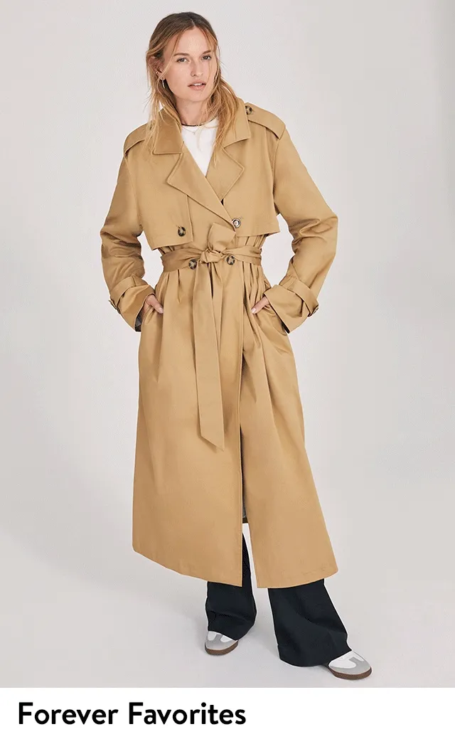 A woman wearing a khaki-colored trench coat with a T-shirt, trousers and sneakers.