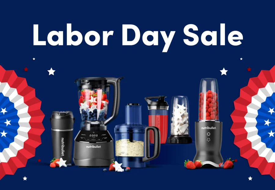 A collection of nutribullet products is arranged against a dark blue background with red, white, and blue fan decorations on the sides. The devices are filled with ingredients like strawberries, blueberries, ice, and smoothie mixes. Small patriotic stars and strawberries are scattered around the display, emphasizing a festive, celebratory theme.