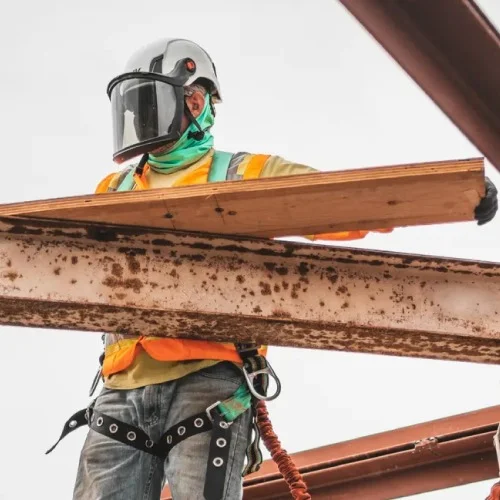 A construction worker in full safety gear, including a helmet with a face shield, safety vest, and harness, maneuvers a wooden plank on a steel beam.