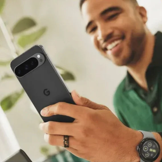 A person smiles while holding a dark gray Google Pixel phone in their left hand. They are also wearing a smartwatch on their left wrist and a ring with lettering on their left ring finger.