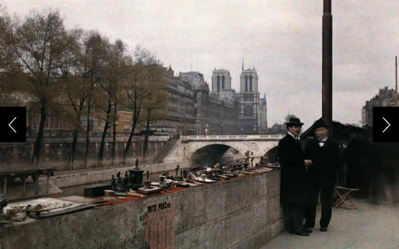 Two men stand next to trinkets for sale on the banks of the Seine in the 1920s with Notre Dame in the background.