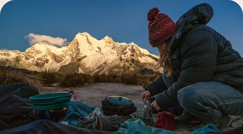 A person sitting on the ground organizes their climbing gear near a snow-covered mountain.