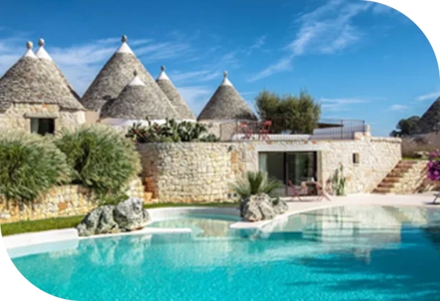 Stone villa with conical roofs and lush greenery surrounding a turquoise pool under clear skies.
