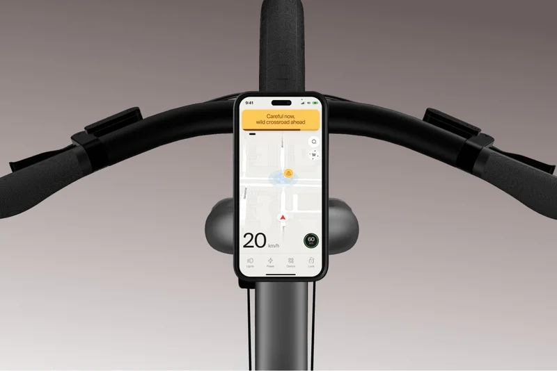 A close-up view of the Cowboy bike’s handlebars with a mounted smartphone displaying a navigation interface. A yellow warning notification reads, “Careful now, wild crossroad ahead,” and the speedometer shows 20 km/h with a green battery indicator at 60%.