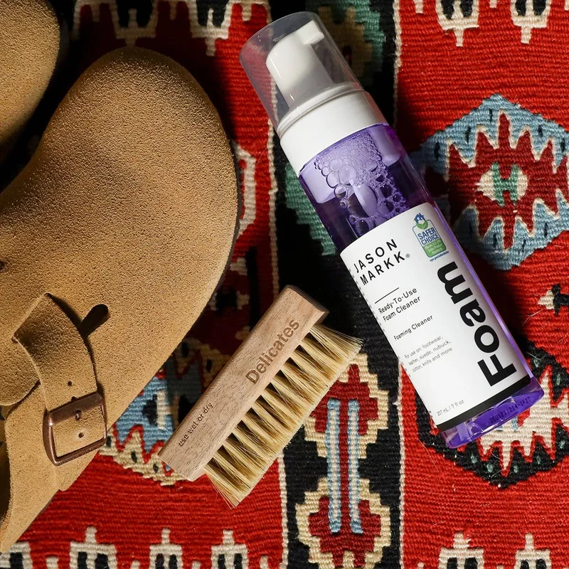 A flat-lay composition shows a tan suede shoe, a wooden brush with bristles labeled 'Delicates use wet or dry', and a bottle of JASON MARKK Ready-To-U...