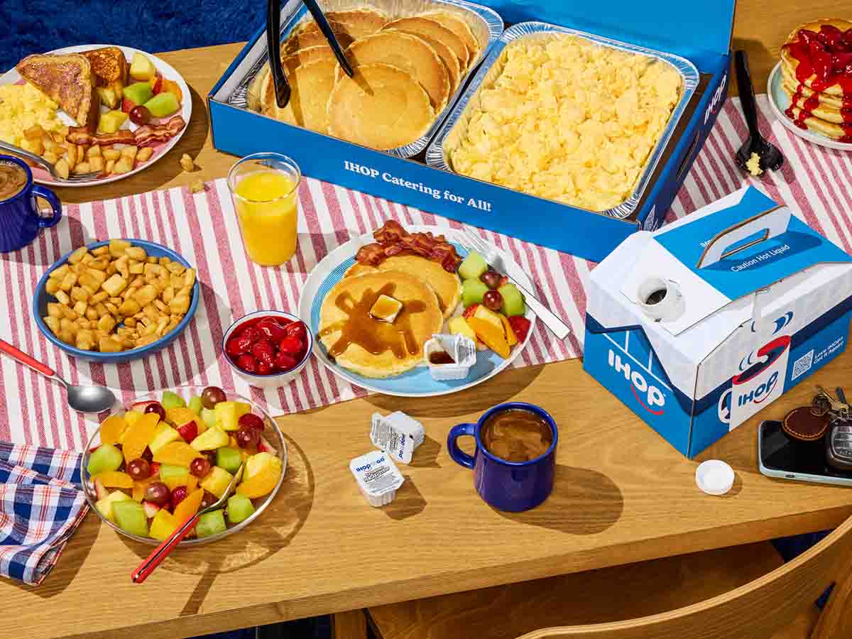 Catering spread with trays of pancakes and eggs, plates of potatoes and fruit.