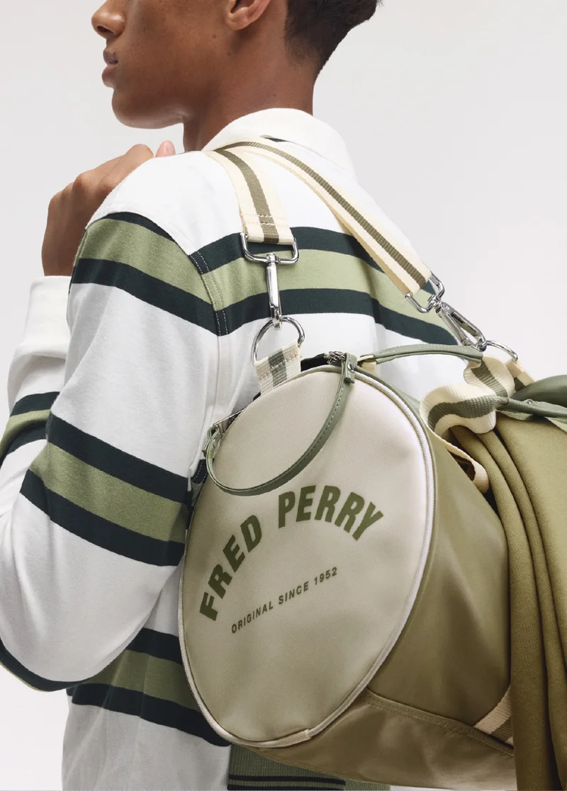 Male model has an olive green and ecru barrel bag with arched Fred Perry branding slung over his left shoulder