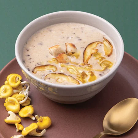 A bowl of creamy mushroom soup with sliced mushrooms and croutons, served on a brown plate with yellow mushrooms and a gold spoon.