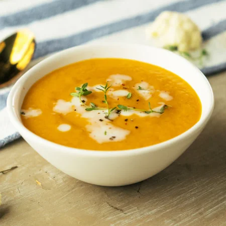 A bowl of orange-colored soup with cream swirls and a sprig of herbs on a wooden table.