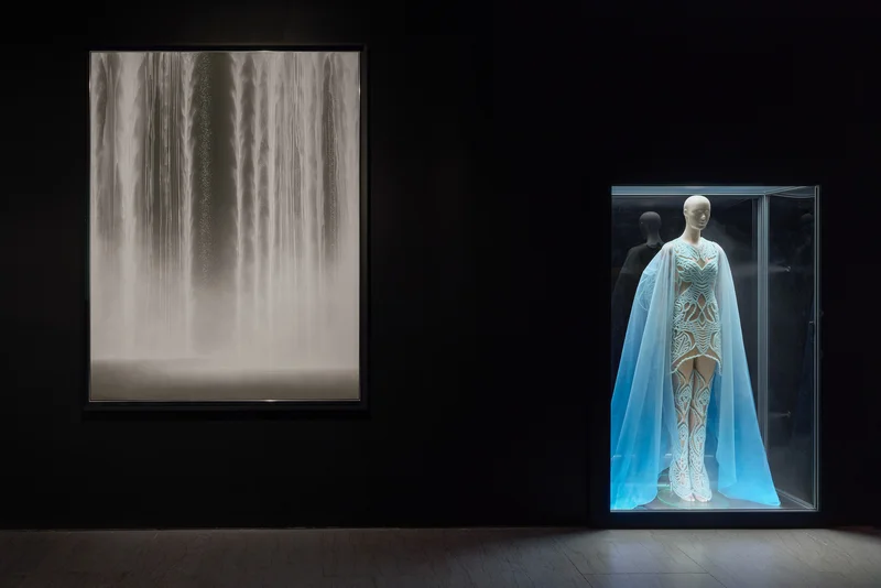 A glowing blue dress on a mannequin next to an artwork that evokes falling water