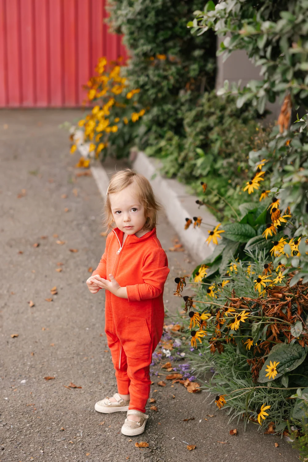 kid wearing red shay romper stood next to yellow flowers
