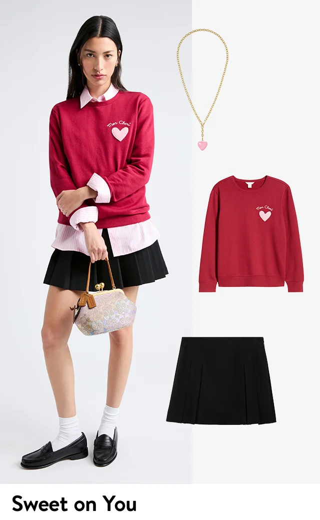 A woman wearing heart-embellished sweater over a button-up with a skirt and handbag. The sweater, skirt and a heart pendant necklace.