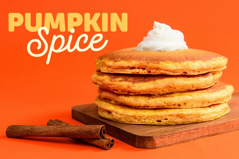 A full stack of Pumpkin Spice Pancakes with whipped topping on a wooden cutting board. Two cinnamon sticks are beside it.