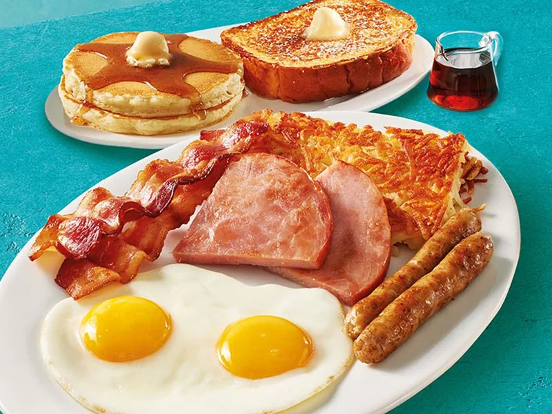 A plate with two eggs, hash browns, two slices of ham, two bacon strips, and two pork sausage links. A second plate has two buttermilk pancakes and a slice of Thick ‘N Fluffy French Toast.