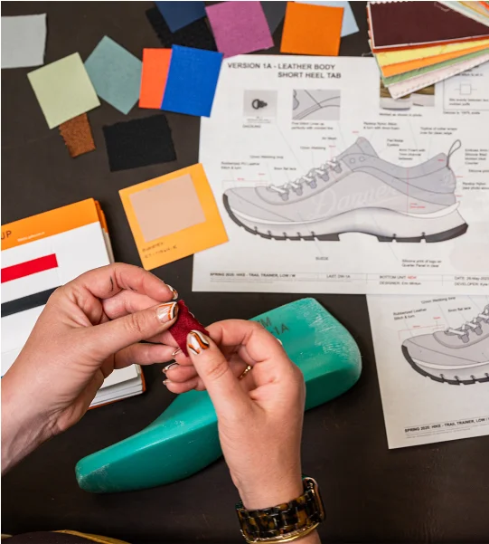 A set of hands holds a color swatch over a desk with boot last, swatches, and technical drawings in the background.