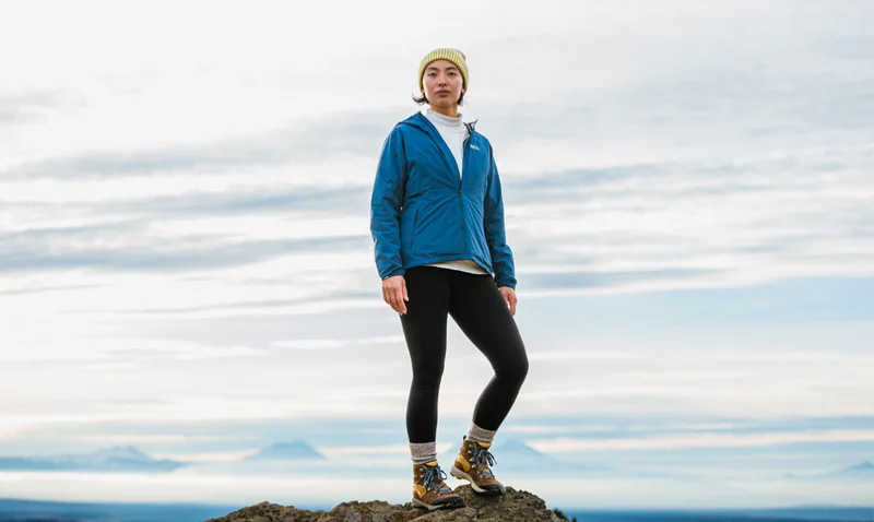 A woman stands on a rocky outcrop under a vast, cloudy sky with distant mountain peaks behind her. She is dressed in a blue jacket, white shirt, black leggings, and hiking boots, topped with an off-white beanie. Her expression is thoughtful as she gazes into the camera.