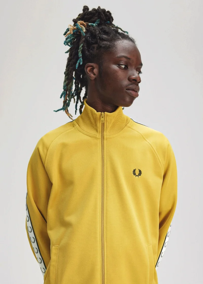 Male Black model with a long twisted hairstyle stands straight and looks pensively to the side while wearing a bright yellow track jacket with white taping and black laurel wreath logos