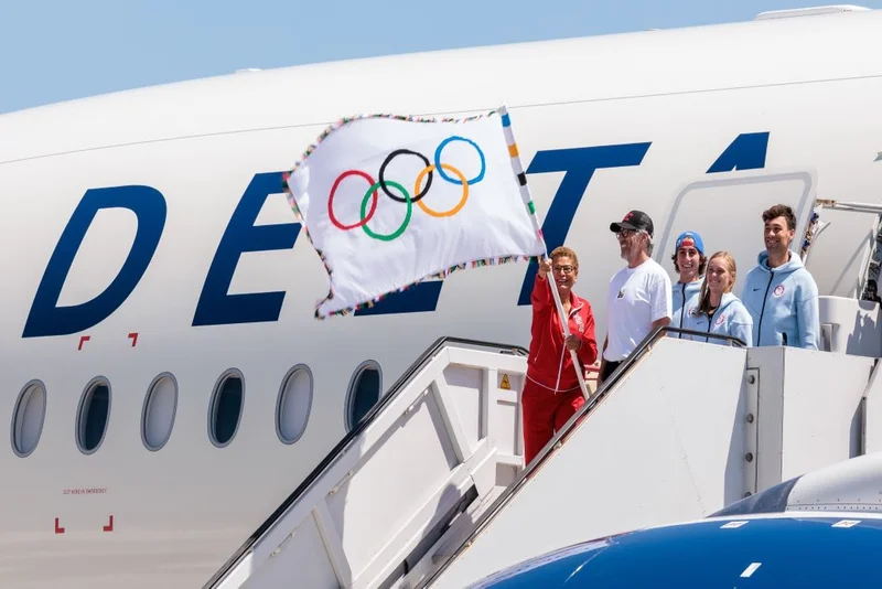 Notable dignitaries deboard the Delta aircraft, outfitted with the LA28 livery. A woman is seen waving the Olympic Flag.