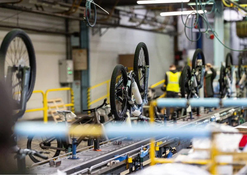 A factory worker assembling a Cowboy bike on a production line, focusing on the rear wheel and drivetrain.
