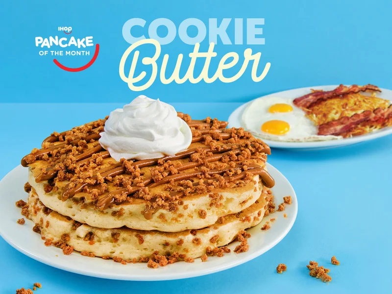 A shot of two Cookie Butter Pancakes, topped with Biscoff® Cookie Butter, cookie pieces, and whipped cream, with a plate of eggs sunny side up, hash browns, and bacon strips in the background.