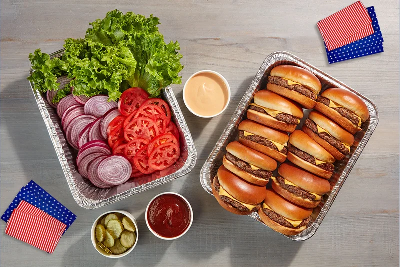 Two catering tins. One filled with IHOP Steakburgers and the other filled with fixins’ like lettuce, tomato, and onions. A cup of pickles, ketchup and spread are also on the table, along with American flag inspired napkins.